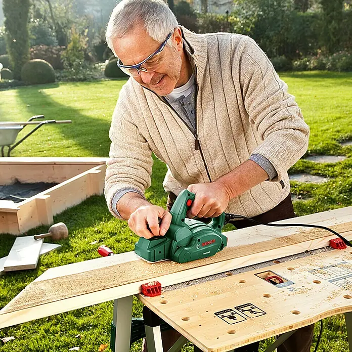 Bosch Cepilladora de mano PHO 1500 - 550 W, Profundidad de cepillo: 0 mm - 1,5 mm, 19.500 r.p.m. - Un hombre usa un cepillo eléctrico Bosch para alisar una tabla de madera, usando gafas de seguridad para protección.