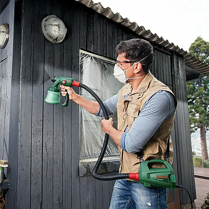 Bosch Sistema de pulverización de pintura PFS 2000 - Apto para: Todos los colores, 440 W, Caudal: 0 l/min - 0,2 l/min, Velocidad de trabajo: 1,5 m²/min - Un hombre usa un sistema de pulverización de pintura Bosch PFS 3000-2 para pintar una casa, con máscara y gafas.