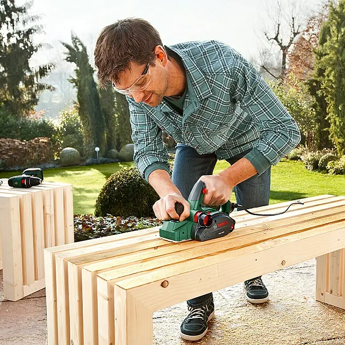 Bosch Cepilladora eléctrica PHO 2000 - 680 W, Profundidad de cepillo: 0 mm - 2 mm - Un hombre usa un cepillo eléctrico Bosch para trabajar una pieza de madera. El cepillo es verde y rojo, mientras que el hombre lleva gafas de seguridad.