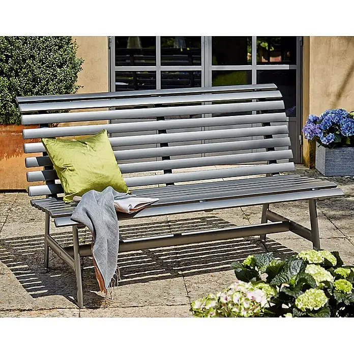 Sunfun Maja Gartenbank - B x T x H: 150 x 72 x 88 cm, Aluminium, Anthrazit - Eine graue Gartenbank mit Kissen und Decke vor einer Hauswand mit Pflanzen und Blumen.