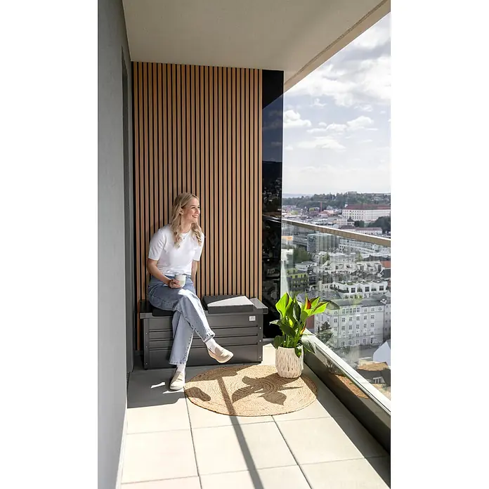 Biohort Kissenbox - 101 x 46 x 46,5 cm, Metall, Anthrazit - Eine Frau sitzt auf einer dunkelgrauen Outdoor-Sitzbank mit Stauraum auf einem modernen Balkon und genießt die Aussicht.