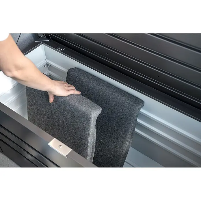 Kissenbox - 101 x 46 x 46,5 cm, Metall, Anthrazit - Eine Hand legt graue Kissen in eine dunkelgraue Outdoor-Aufbewahrungsbox mit weißem Innenraum.