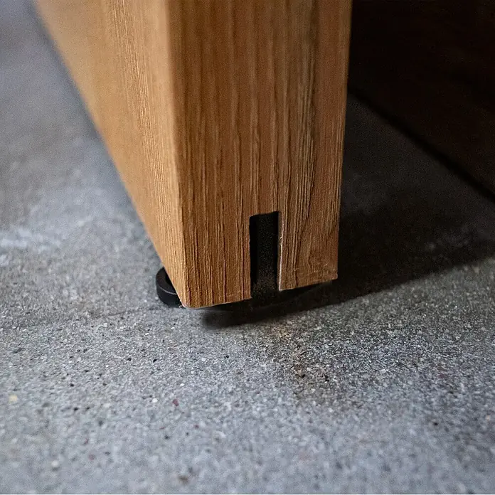 Inova Schiebetür-Komplettset Holz Scheunentor Eiche inkl. Softclose - 775 x 2.065 mm - Nahaufnahme eines hellbraunen Holzmöbelfußes mit schwarzem, rundem Bodengleiter auf Betonboden.