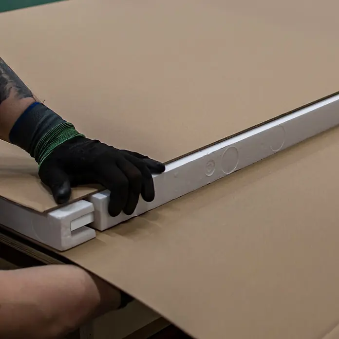 Inova Schiebetür-Komplettset Holz Weiß mit Beschlag und Bügelgriff in schwarz -  - Behandschuhte Hand platziert weiße Schaumstoffverpackungsecken auf braunem Karton. Verriegelnder Schaum sichert das Produkt.