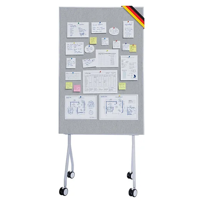 Pinnwand Präsentationstafel Giant - 120 x 80 cm, Filz, Grau - Eine graue Filz-Pinnwand auf Rollen mit weißen Beinen, bedeckt mit Dokumenten, Notizen und Bauplänen, und einem kleinen Deutschlandflaggenband.