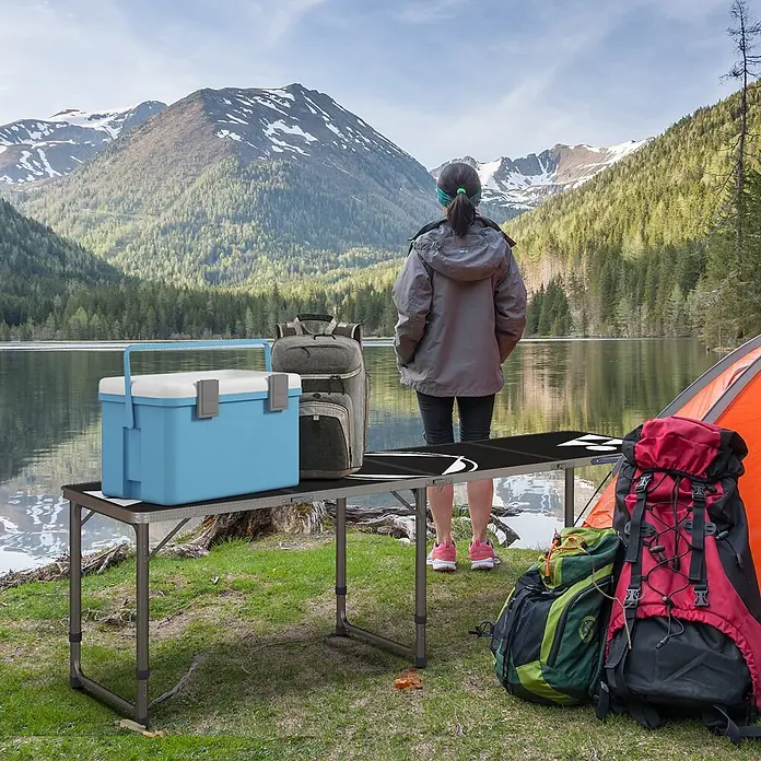 Outsunny Campingtisch - Aluminium, MDF, Schwarz, Klappbar - Ein dunkelgrauer Klapptisch mit Metallbeinen, darauf eine blaue Kühlbox und ein Rucksack, an einem See mit Bergen.