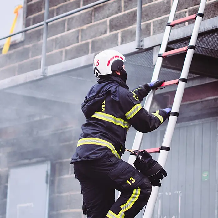 Telesteps Teleskopleiter Rescue Line Feuerwehr- & Rettungsleiter 4,1m – Teleskopleiter für 3 Pers - Anzahl Tritte: 13 Stk., Arbeitshöhe: 5.000 m, Belastbarkeit: 500 kg, Aluminium - Feuerwehrmann klettert eine weiße Ausziehleiter mit roten Sprossen an einem Gebäude hoch.