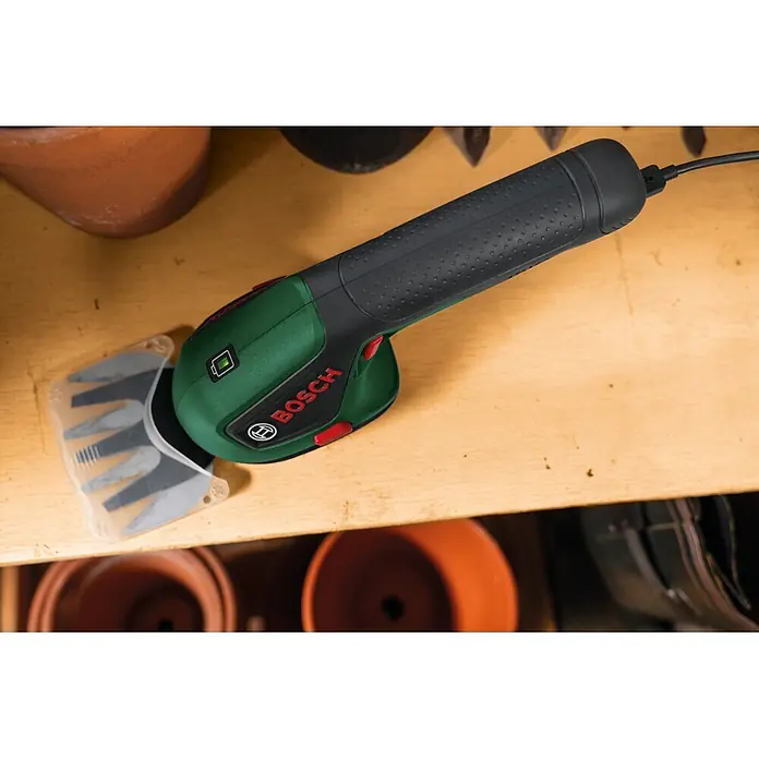 Bosch Akku-Grasschere ISIO 4 - 4 V, 1 Akku - Grüne und schwarze Bosch Akku-Strauchschere mit transparentem Klingenschutz, grünem Akku-Symbol, auf einem Holzregal.