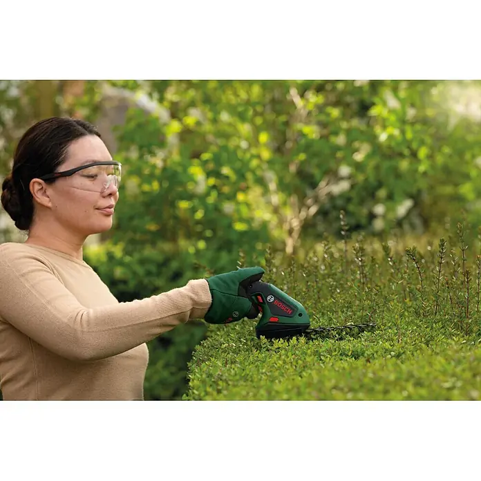 Bosch Akku-Gras- & Strauchschere ISIO 4 - 4 V, 1 Akku - Frau in Schutzbrille und grünen Handschuhen trimmt eine Hecke mit einer grün-schwarzen Bosch Strauchschere.