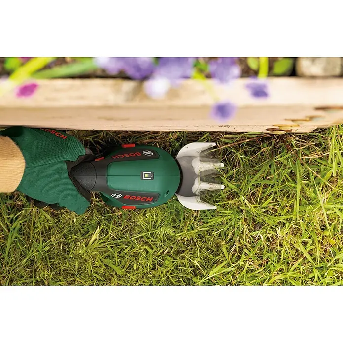 Bosch Akku-Gras- & Strauchschere ISIO 4 - 4 V, 1 Akku - Grüne Bosch Grasschere trimmt Rasenkante an Holzrand, gehalten von behandschuhter Hand.