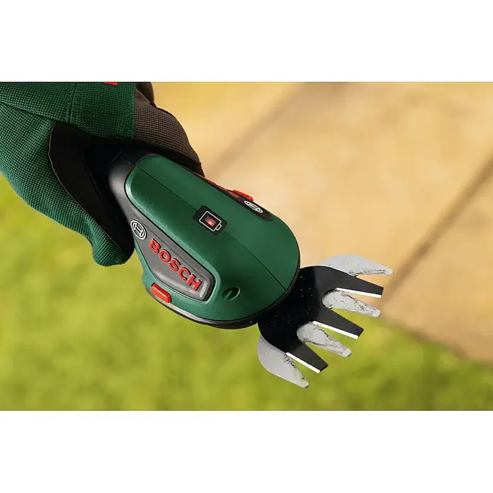 Bosch Akku-Gras- & Strauchschere ISIO 4 - 4 V, 1 Akku - Eine behandschuhte Hand hält eine grün-schwarze Bosch Grasschere mit roten Akzenten, bereit zum Gartenschnitt.