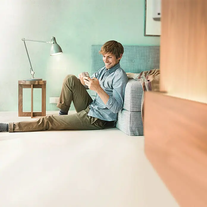 Osram Lightify Bombilla LED Classic A60 - E27, Intensidad regulable, Blanco neutro, 310 lm, 9,5 W - Un hombre está sentado en el suelo usando su teléfono inteligente. Una lámpara y una mesa auxiliar están junto a él. La habitación es luminosa y moderna.