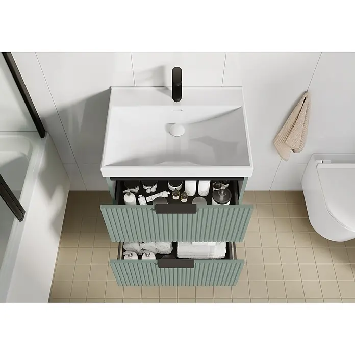 Riva Yasumi Waschtischunterschrank - B x T x H: 60 x 45,2 x 63,5 cm, Sage, Matt - Ansicht von oben auf einen modernen Badezimmerschrank mit weißem Waschbecken und schwarzem Wasserhahn. Die hellgrünen, geriffelten Schubladen zeigen Toilettenartikel und weiße Handtücher.