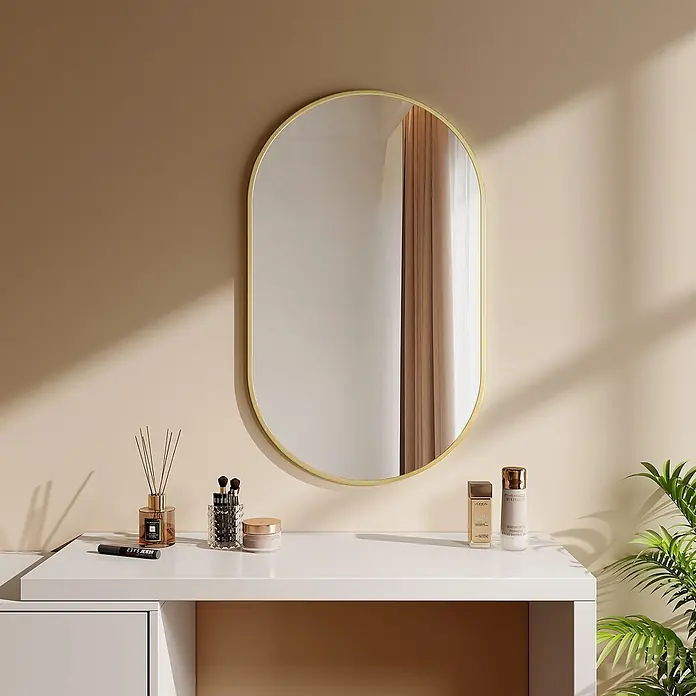 Emke Rahmenspiegel OM18 - 100 x 60 cm, Glas - Ein ovaler Spiegel mit Goldrahmen über einem weißen Schminktisch mit Beauty-Produkten und einer Pflanze, der warmes Licht reflektiert.