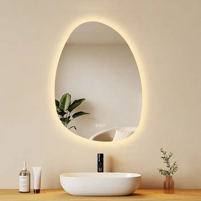 Emke Lichtspiegel Glowrim LM34 - 75 x 55 cm - Unregelmäßiger LED-Spiegel über weißem Waschbecken, der Pflanze und Sessel widerspiegelt.