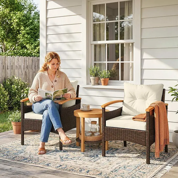 Outsunny Loungemöbel-Set - 3 -tlg., PE-Rattan, Stahl, Polyester, Akazienholz, Schaumstoff, PP-Baumwolle, inkl. Kissen, Braun, Cremeweiß, Naturholz - Zwei braune Korbsessel mit cremefarbenen Kissen und ein runder Holztisch im Freien, auf einer Holzterrasse mit einer sitzenden Person.