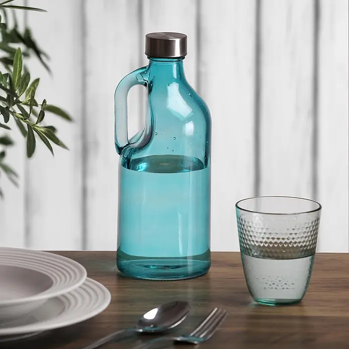Versa Botella con asa - 1,1 l, Vidrio, Azul - Botella de agua de cristal azul con tapa metálica y vaso texturizado a juego sobre mesa de madera con vajilla.