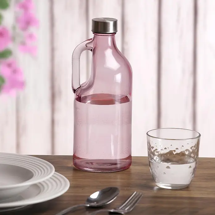 Versa Botella con asa - 1,1 l, Vidrio, Rosa - Botella de agua de cristal rosa con tapa plateada y asa integrada, llena de agua, sobre una mesa de madera con un vaso.