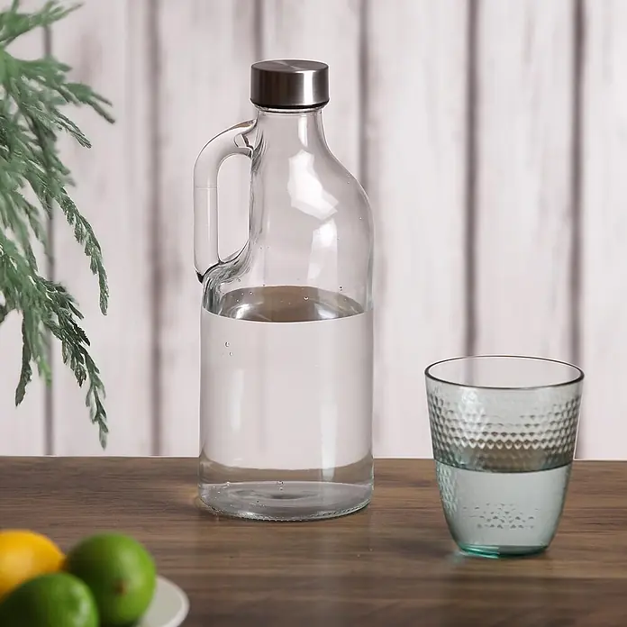 Versa Botella con asa - 1,1 l, Vidrio, Incoloro - Jarra de vidrio transparente con asa y tapa metálica, llena de agua, junto a un vaso en una mesa de madera.