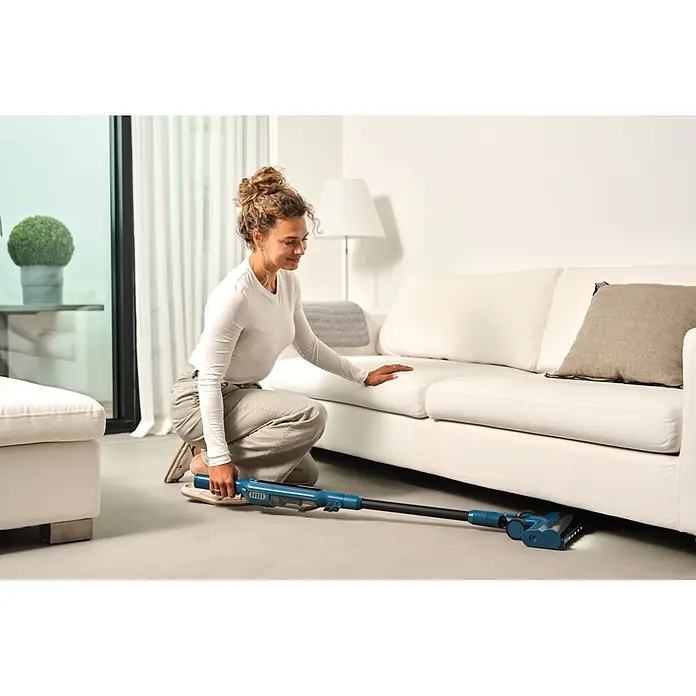 Nilfisk Aspirador de batería Move - 150 W, Volumen de recipiente: 0,2 l, Negro, Con soporte de pared - Mujer usando una aspiradora escoba azul y negra para limpiar debajo de un sofá blanco.