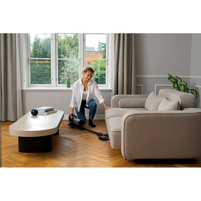 Nilfisk Aspirador de batería S1 Pet Care - 29,6 V, 1 batería, Volumen de recipiente: 0,7 l - Una mujer usa una aspiradora escoba negra y plateada para limpiar el suelo de madera bajo un sofá.