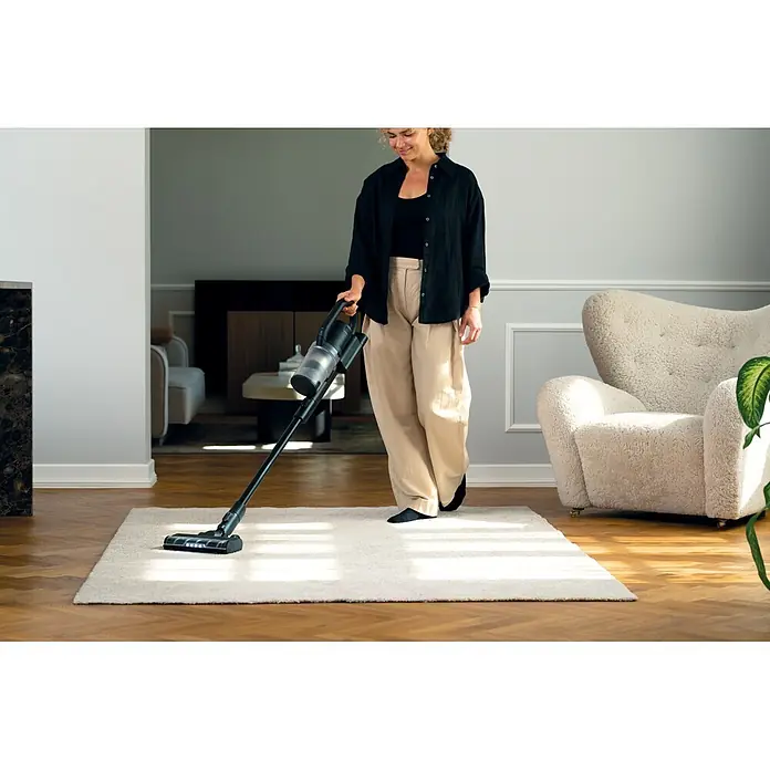 Nilfisk Aspirador de batería S1 Pet Care - 29,6 V, 1 batería, Volumen de recipiente: 0,7 l - Una persona aspira una alfombra clara con una aspiradora de escoba inalámbrica negra y gris.