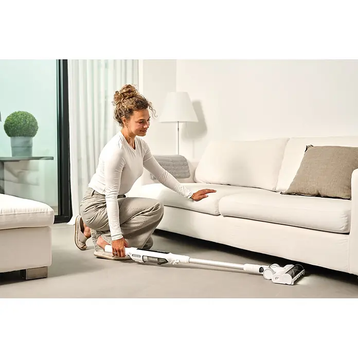 Nilfisk Aspirador de batería Move - 150 W, Volumen de recipiente: 0,2 l, Blanco, Con soporte de pared - Aspirador escoba inalámbrico blanco limpiando debajo de un sofá, demostrando su alcance.