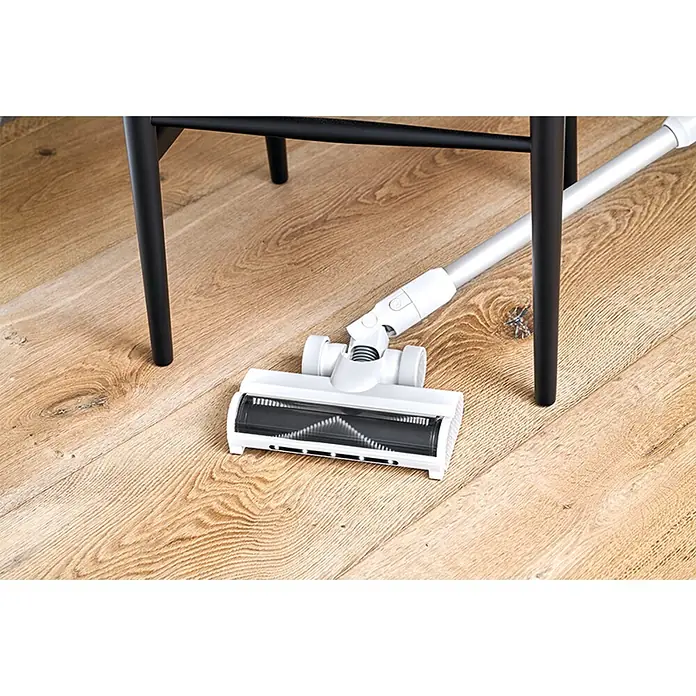 Nilfisk Aspirador de batería Move - 150 W, Volumen de recipiente: 0,2 l, Blanco, Con soporte de pared - Cabezal de aspiradora blanco con cepillo giratorio negro sobre suelo de madera, con mango plateado junto a patas de mueble negras.