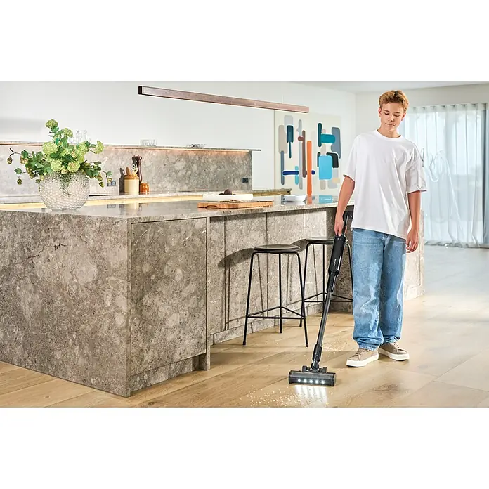 Nilfisk Aspirador de batería Move - 150 W, Volumen de recipiente: 0,2 l, Con base de carga - Un aspirador de escoba negro limpia la suciedad de un suelo de cocina de madera.