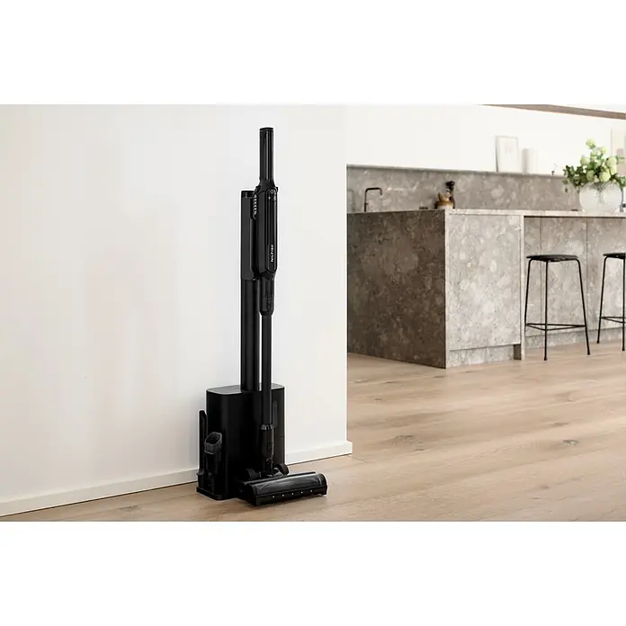 Nilfisk Aspirador de batería Move - 150 W, Volumen de recipiente: 0,2 l, Con base de carga - Aspiradora de escoba Nilfisk negra en su base con accesorios, sobre suelo de madera en cocina.
