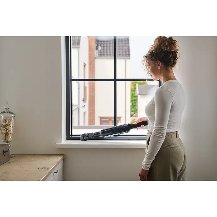 Nilfisk Aspirador de batería Move - 150 W, Volumen de recipiente: 0,2 l, Con base de carga - Mujer limpiando un alféizar con un aspirador de mano Nilfisk negro.