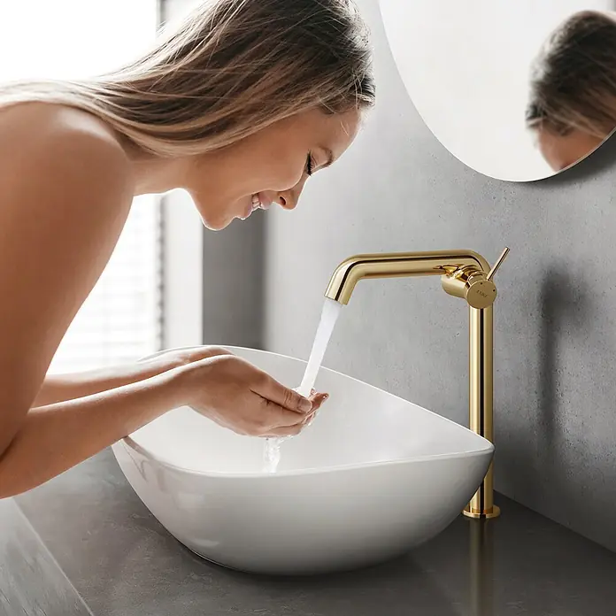 Emke Waschtischarmatur Flowcurv XF24 - Gold, Glänzend - Frau wäscht ihr Gesicht mit Wasser aus einem stilvollen goldenen Badarmatur, passend zu einem modernen weißen Waschbecken.