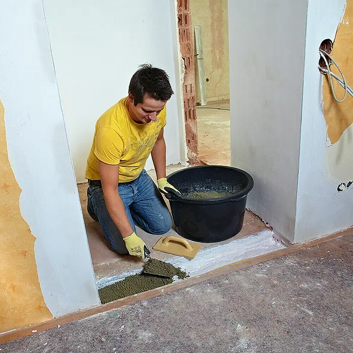 Quick-Mix Schnellestrich - 30 kg, Schichtdicke: 60 mm - Ein Mann kniet und schaufelt Estrichmasse vom Boden, während er eine Kelle und einen Eimer verwendet, um den Raum vorzubereiten.