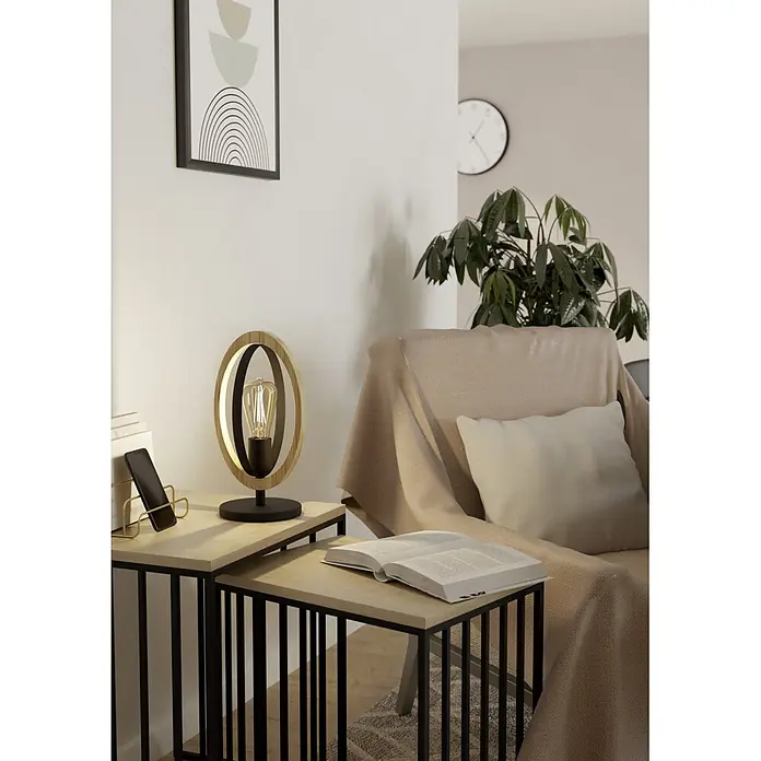 Eglo Basildon Tischleuchte - 40 W, L x B x H: 19 x 16 x 34,5 cm, E27 - Beleuchtete Tischlampe mit Holz- und schwarzen Metallkreisen auf einem hellen Holztisch neben einem Sessel.