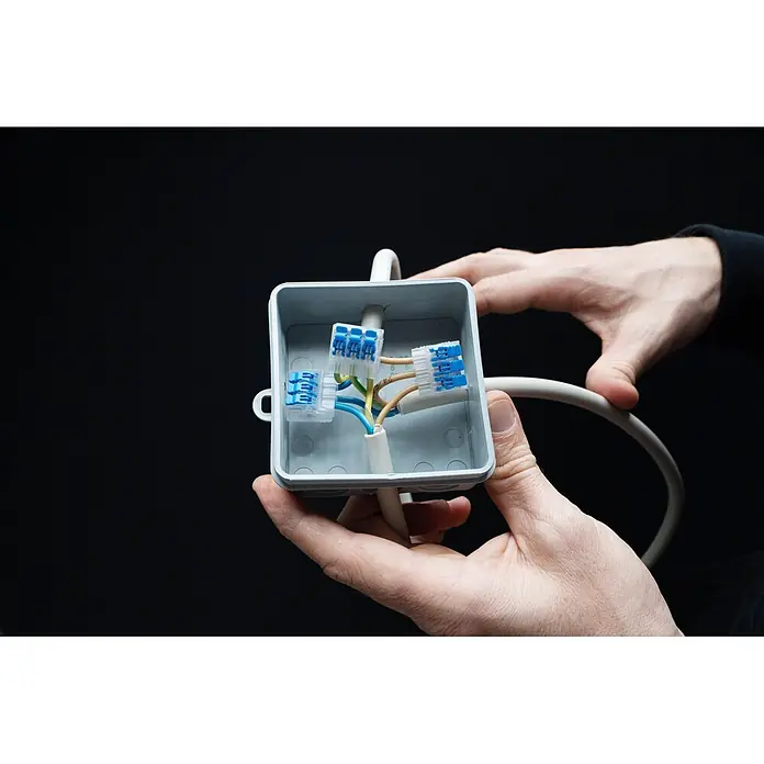 Ideal Hebelklemme 2/3/5-fach Gen II Kombipack - 30 Stk. - Hände halten grauen Abzweigkasten mit blauen Kabelverbindern und farbigen Drähten.
