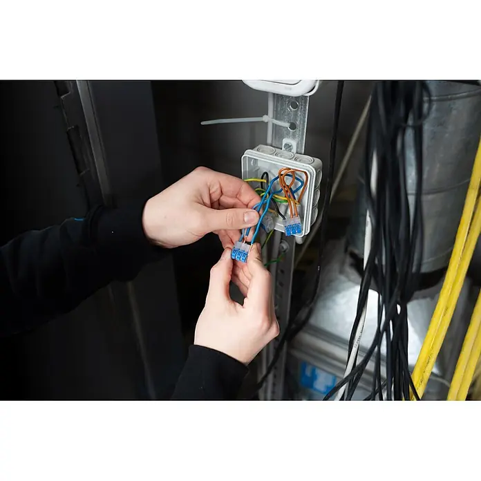Ideal Hebelklemme 2/3/5-fach Gen II Kombipack - 30 Stk. - Hände verbinden blaue und orange elektrische Kabel mit Hebelklemmen in einer weißen Abzweigdose bei der Elektroinstallation.