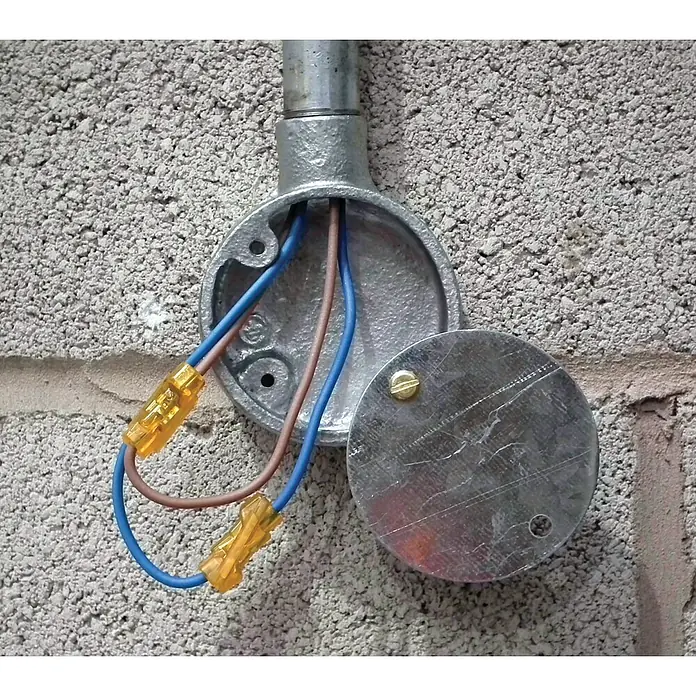 Ideal Stoßverbinder Drahtverbinder Spliceline 30-1042-10 - 10 Stk. - Runde elektrische Metallabzweigdose mit Rohrleitung, offen, zeigt blaue und braune Kabel, die mit gelben Klemmen an einer Betonwand verbunden sind.