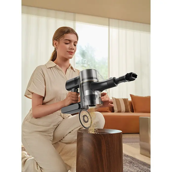 Mova Aspirador de batería S5 Sense - 570 W, Volumen de recipiente: 0,6 l - Aspiradora de mano negra y plateada con depósito transparente, siendo vaciada por una mujer en un cubo de madera.