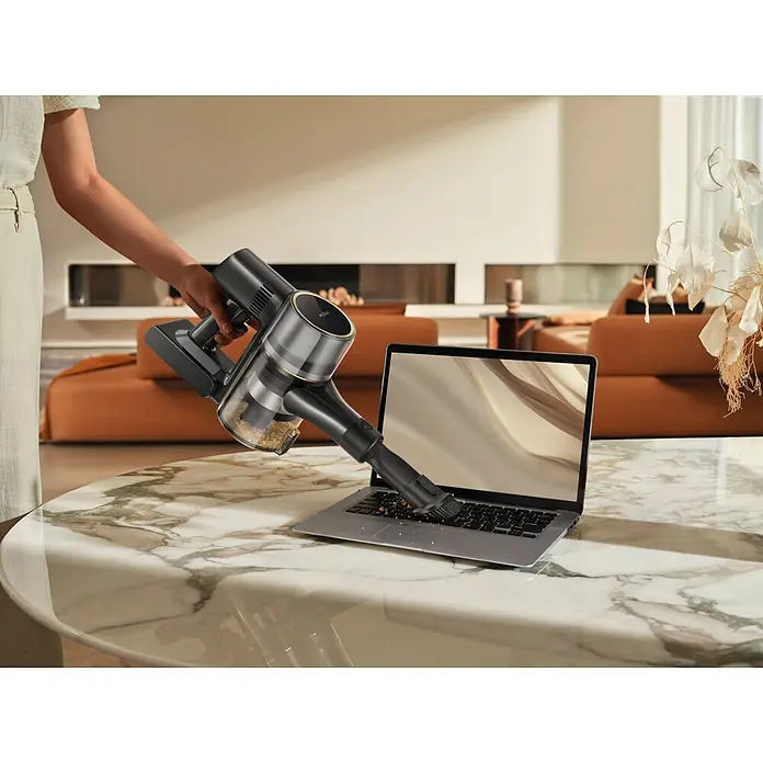 Mova Aspirador de batería S5 Sense - 570 W, Volumen de recipiente: 0,6 l - Aspirador inalámbrico gris oscuro con detalles dorados limpiando migas de un teclado de portátil sobre una mesa de mármol.