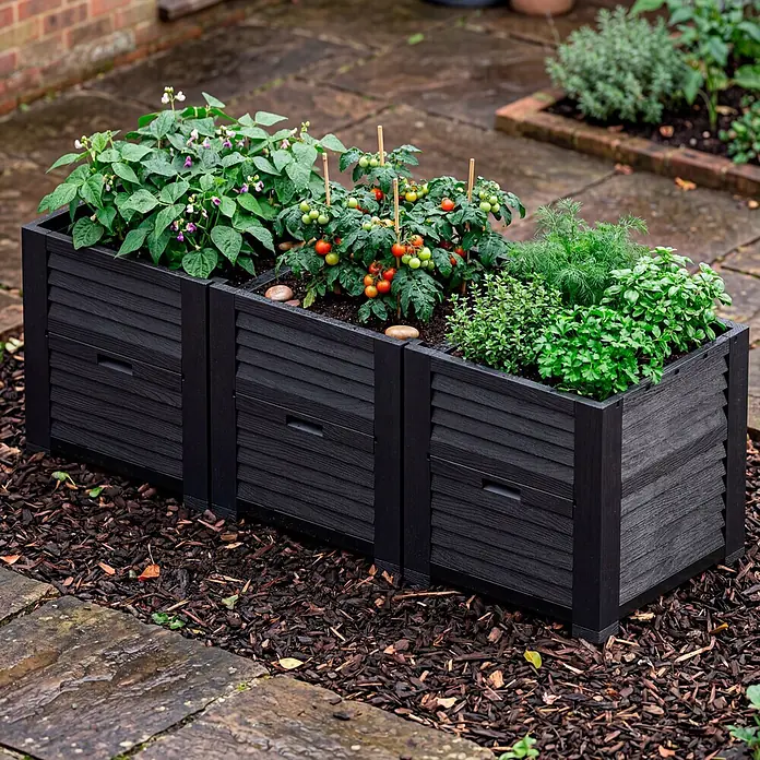 Hochbeet mit 3 Staufächern - 225 x 75 x 78 cm, Kunststoff - Drei schwarze Lamellen-Hochbeete mit Pflanzen auf einer Terrasse mit Mulch.