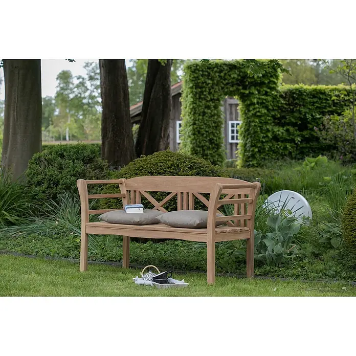 Bene Living Gartenbank Föhr - B x T x H: 130 x 53 x 83 cm, Teakholz, Natur - Teak-Gartenbank mit gekreuzter Rückenlehne, zwei grauen Kissen und einem Buch in einem üppigen Garten.