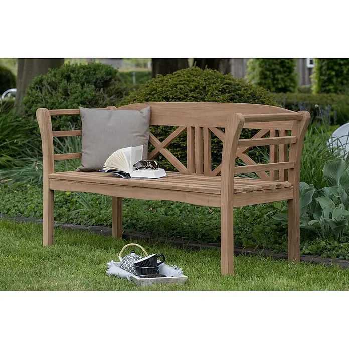 Bene Living Gartenbank Föhr - B x T x H: 130 x 53 x 83 cm, Teakholz, Natur - Gartenbank aus Naturholz mit grauem Kissen, offenem Buch und Sonnenbrille.