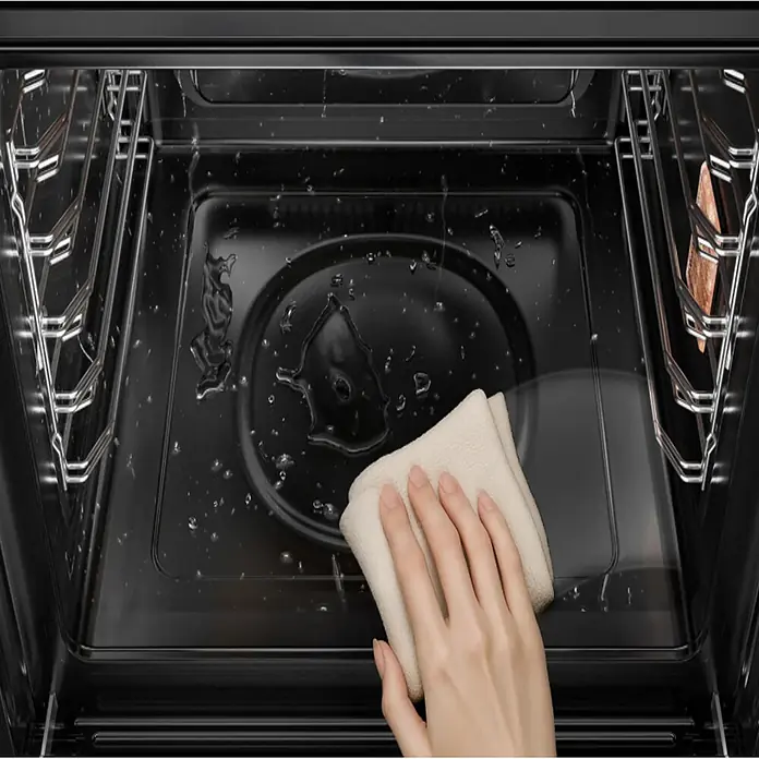 Midea Horno empotrado MBOG2-D2M90 - Volumen horno: 70 l, Número de tipos de calentamiento: 9 ud. - Mano limpiando agua del interior de un horno negro con un paño claro, mostrando parrillas metálicas.