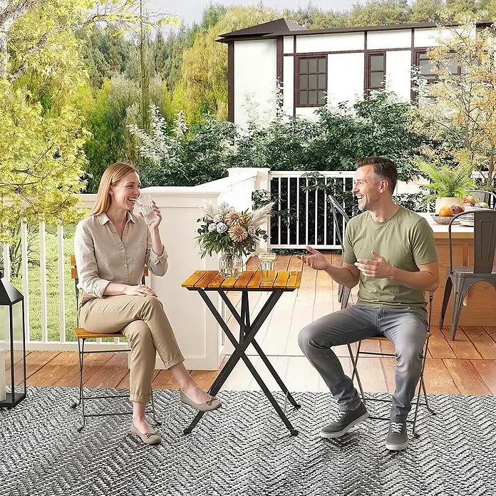 Outsunny Gartentisch-Set - Gesamtumfang: 3 -tlg., Stahl, Akazienholz, Klappbar - Paar trinkt an einem kleinen Bistrotisch aus Holz und Metall mit passenden Stühlen auf einem gemusterten Outdoor-Teppich.