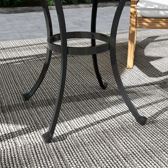 Outsunny Garten-Beistelltisch rund - Aluminiumguss, Schwarz - Schwarzes Metall-Outdoor-Tischgestell mit geschwungenen Beinen auf einem gemusterten grau-weißen Teppich, neben einem Holzstuhl.