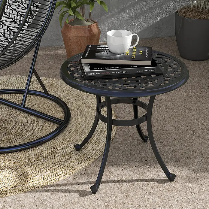 Outsunny Garten-Beistelltisch rund - Aluminiumguss, Schwarz - Schwarzer Gusseisen-Beistelltisch für den Außenbereich mit verziertem Oberteil, Tasse und Büchern darauf, neben einem Hängesessel auf einer Terrasse.
