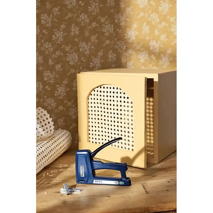 Rapid Grapadora manual RPC4 - Profundidad de grapado: 6 mm - 16 mm, Apto para: Papel - Grapadora Rapid RPC4 azul sobre superficie de madera junto a un mueble con rejilla de caña y un rollo de rejilla de caña.