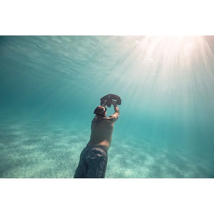 Sublue Tauchscooter Navbow 2026 - L x B x H: 486 x 327 x 177 mm - Mann unter Wasser hält einen schwarzen Tauchpropeller zur Oberfläche, mit Sonnenstrahlen, die durch das Wasser dringen.