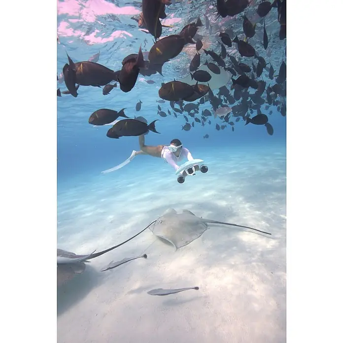 Sublue Tauchscooter Mix 2026 - L x B x H: 465 x 230 x 230 mm - Person mit Flossen und Schnorchel nutzt einen hellblauen Unterwasserscooter inmitten von Fischen und Rochen.
