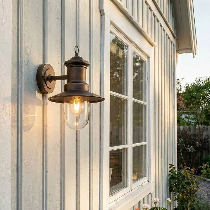 Lindby Außenwandleuchte Eloretta - messing / gold, messing / gold, IP44 - Bronze Außenwandleuchte mit klarem Glasschirm und sichtbarer Glühbirne, montiert an einem hellen Holzhaus neben einem Fenster.